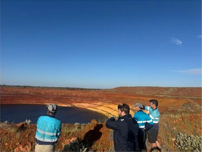 Four people standing beside the water-filled Western Queen South pit in a red-earth mining landscape under a clear blue sky.