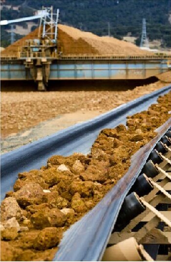 Conveyor carrying ore beside a large stockpile at an industrial minerals operation, with plant equipment and forested hills behind.