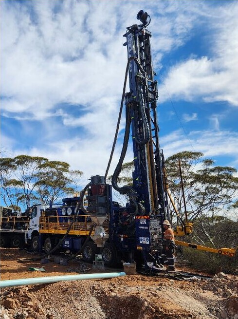 Drilling rig and support truck operating beside native vegetation under a blue, cloudy sky.