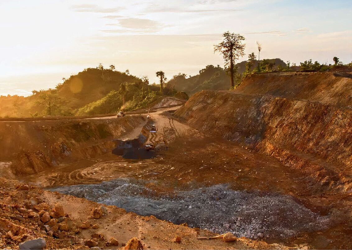 Open-cut mining landscape with a broad dirt haul road, excavated slopes, exposed rock and tropical vegetation in the background.