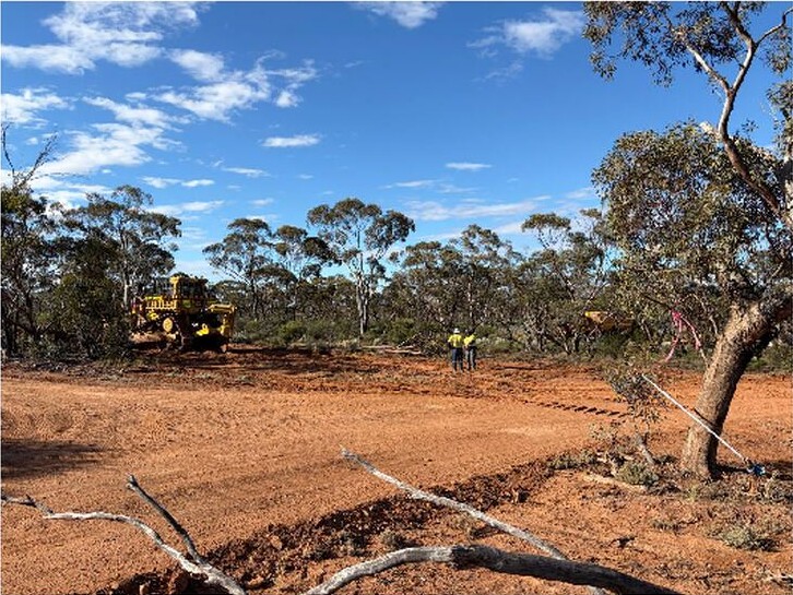 Cleared red-earth area bordered by bushland, with heavy machinery and workers visible beneath a blue sky.