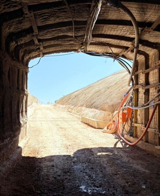 View from inside a reinforced mine tunnel looking out toward a sunlit open haulage route, with cables and equipment along the tunnel walls.