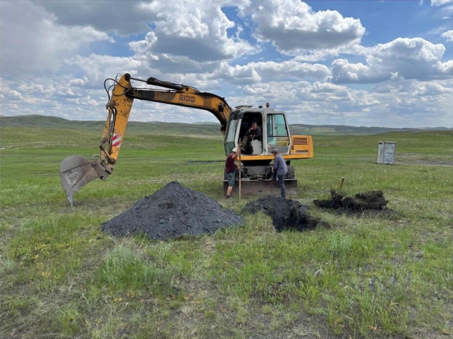 Excavator beside an exploration test pit, with people, excavated dark material and grassy hills under a cloudy sky.