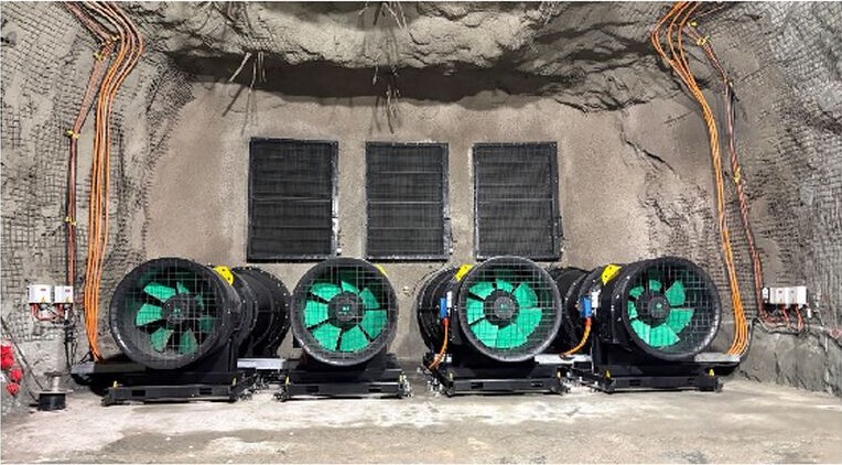 Four large interim ventilation fans installed underground in a rock-walled mine chamber, with power cables and ventilation grilles behind them.