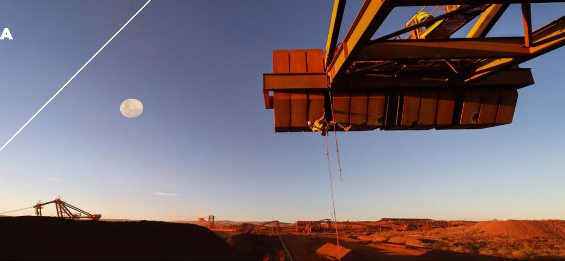 Open-pit mining landscape at dusk, with large industrial mining machinery silhouetted against the sky and a moon above the site.